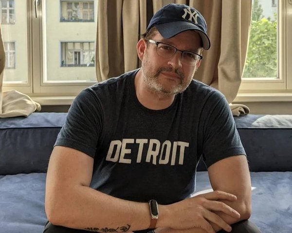 Sascha Pallenberg sitzt in einem Zimmer, trägt ein 'Detroit'-Shirt und eine Brille mit New York Yankees Cap.