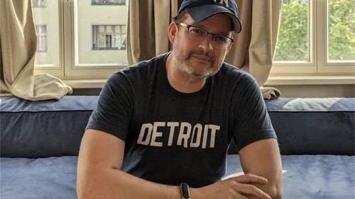 Sascha Pallenberg sitzt in einem Zimmer, trägt ein 'Detroit'-Shirt und eine Brille mit New York Yankees Cap.