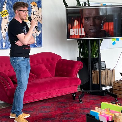 Matthias Krahé präsentiert vor einem Monitor auf dem Arnold Schwarzenegger und der Text BULL gezeigt wird