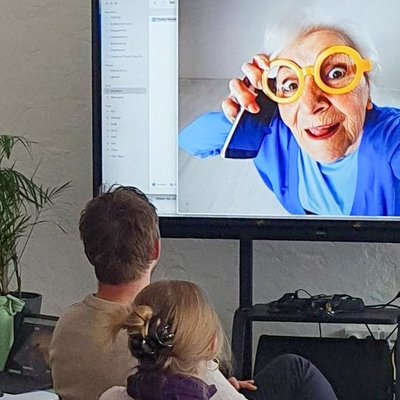 Teilnehmer schauen sich ein Bild einer älteren Frau mit gelber Brille und Smartphone auf einem Bildschirm an.