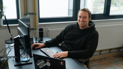 Roman Lohfink in einem schwarzen Hoodie sitzt lächelnd vor einem Computer in einem Büro.