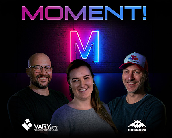 Lars Michaelis, Sarah Tavčer und Matthias Rosa, lächelnd vor einer pink-blau beleuchteten Neonwand mit großem „M“. Logos von VARY.FY und robotspaceship sowie der Schriftzug „MOMENT!“ sind zu sehen.